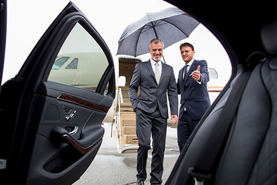 Ein Chauffeur hält einen Regenschirm für einen Fahrgast und hilft ihm beim Einsteigen in eine Limousine.